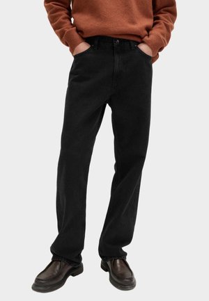 Persona che indossa jeans neri a gamba dritta, mocassini marroni e un maglione color ruggine, in piedi con le mani nelle tasche.