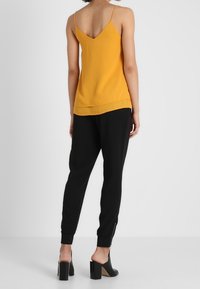 Žlutý tílko bez rukávů s výstřihem do V a rovný lem spojený s černými tepláky s detaily na zipy a zúženým střihem. Boty s blokovým podpatkem.