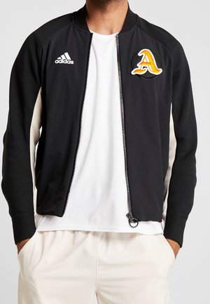 Mężczyzna ubrany w czarną kurtkę Adidas na suwak z białymi bocznymi panelami, białą koszulkę i jasne spodnie z rękami w kieszeniach.
