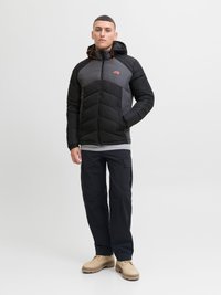 Schwarze und graue Steppjacke mit Kapuze, Reißverschluss und orangefarbigen Akzenten. Getragen mit dunklen Hosen und beigen Schuhen. Glatte Textur, gepolstertes Design.