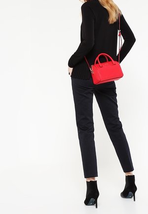 Femme portant un haut noir à manches longues, un pantalon foncé, des bottines noires à talons hauts, portant un petit sac à main rouge avec une bandoulière.