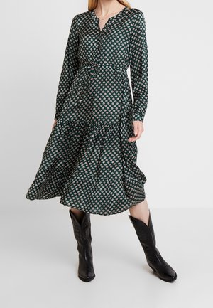 Femme portant une robe midi vert foncé avec un petit motif circulaire blanc, manches longues, et des bottes de cow-boy noires mi-mollet, debout sur un sol blanc.