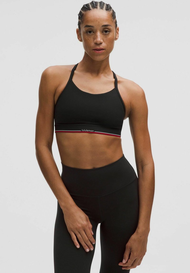 Sujetador deportivo negro con tirantes ajustables, que presenta un logo de Lululemon y una banda horizontal contrastante en rojo y blanco en la parte inferior.