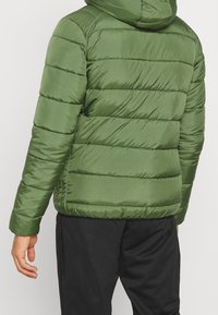 Zelená péřová bunda s kapucí, s vertikálním prošíváním, hladkým povrchem a elastickými manžetami, zkombinována s černými kalhotami.