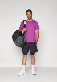 Lilla Nike t-skjorte, svarte treningsshorts og hvite joggesko. Bærer en svart duffelbag med en hvit logostropp. Enkelt, moderne design.