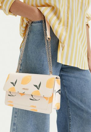 Mano sosteniendo un bolso blanco con estampado de limones amarillos y cadena dorada junto a unos vaqueros azules y una camisa a rayas amarillas.