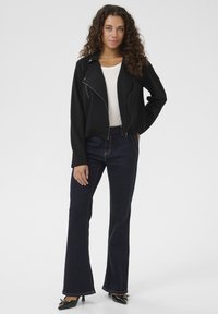 Svart kort jacka med dragkedja och asymmetrisk krage, kombinerad med mörkblå utsvängda jeans och svarta spetsiga klackskor.