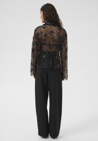 Jachetă din dantelă neagră cu detalii florale din paiete, mânecile lungi și guler, asortată cu pantaloni negri cu talie înaltă.