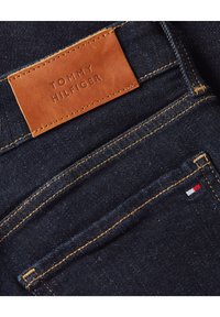 Vaqueros de denim azul oscuro con costuras naranjas, una etiqueta de cuero marrón de Tommy Hilfiger y una pequeña bandera roja, blanca y azul en el bolsillo.
