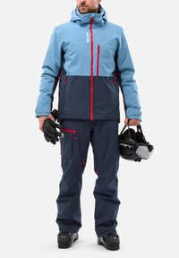 Ski jacket in light blue and navy, featuring red zippers. Paired with navy pants, black gloves, and holding a helmet. Waterproof design.