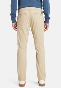 Chino beige con taglio dritto, dotati di due tasche posteriori e chiusure con bottone. Tessuto di cotone liscio con una leggera lucentezza. Sneakers bianche visibili.