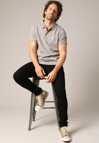 Grijze poloshirt met korte mouwen, zwarte jeans en tan sneakers met witte accenten. Het model zit op een metalen kruk tegen een neutrale achtergrond.