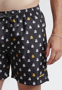 Bañador negro hecho de tela suave, con un patrón repetido de siluetas de stormtroopers en blanco y amarillo, cinturilla elástica.