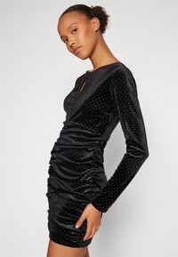 Rochie din catifea neagră cu mâneci lungi, împodobită cu cuie argintii. Prezintă un design plisat și un decolteu în formă de V cu un detaliu în formă de cheie.