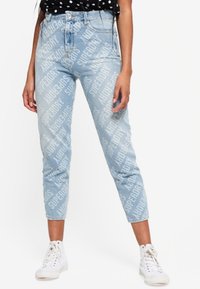 Jean taille haute bleu clair avec un motif répétitif de branding blanc "Superdry", coupe slim, longueur cropped et ourlets retroussés, associés à des baskets blanches.