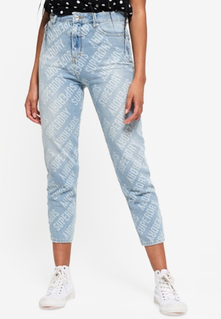 Jean taille haute bleu clair avec un motif répétitif de branding blanc "Superdry", coupe slim, longueur cropped et ourlets retroussés, associés à des baskets blanches.