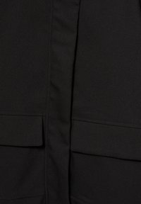 Gros plan sur un tissu noir avec une couture verticale et deux poches horizontales à rabat, une de chaque côté.
