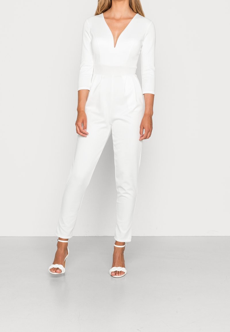 Jumpsuit blanc avec un décolleté en V profond, des manches trois-quarts, une taille cintrée et des jambes fuselées. Porté avec des sandales à talons blanches. Texture lisse.