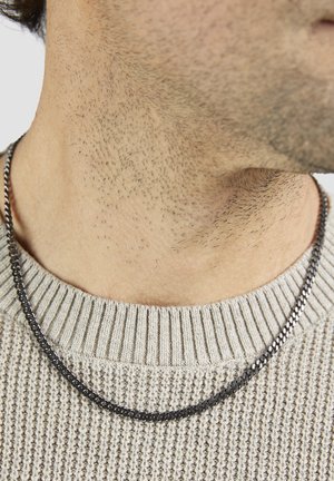 Halskette aus schwarzer Metallkette mit glatter Textur; getragen über einem hellen, gerippten Pullover in neutralem Farbton.