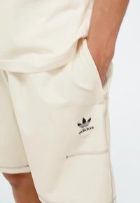 Smėlio spalvos sportiniai šortai, pagaminti iš lygaus audinio, su juodu Adidas logotipu, šoninėmis kišenėmis ir kontrastingomis siūlėmis.