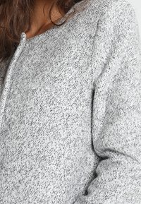 Primo piano di una persona che indossa un morbido pullover grigio a maglia con maniche lunghe e scollo rotondo.