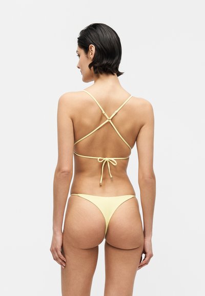 Γυναίκα με κοντά σκουρόχρωμα μαλλιά που φοράει ένα παστέλ κίτρινο μπικίνι με σταυρωτούς ιμάντες και δεμένη φιόγκο στην πλάτη, εμφανιζόμενη από πίσω.