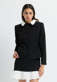 Ung kvinna klädd i en svart knappad jacka med en vit rundad krage och en svart kjol med en vit volangkant.