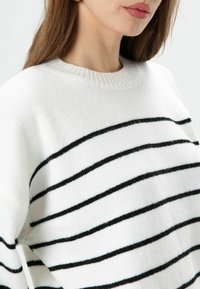 Witte trui met zwarte horizontale strepen, geribde hals en zachte textuur. Heeft een ontspannen pasvorm en een ronde halslijn.