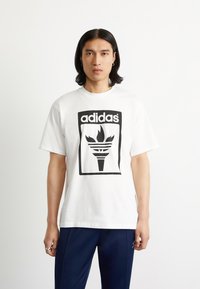 White cotton t-shirt featuring a black adidas logo with a stylized torch design. Short sleeves, crew neck, relaxed fit.