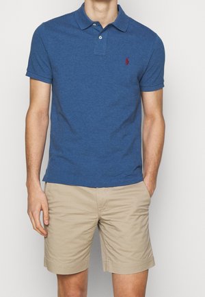 Niebieska bawełniana koszulka polo z kołnierzykiem i krótkimi rękawami. Posiada małe czerwone logo na lewej piersi. Noszona z beżowymi szortami.