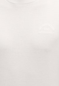 Λευκό ύφασμα με ανάγλυφο κείμενο που αναγράφει «Karl Lagerfeld Paris 21 Rue St Guillaume» στην πάνω περιοχή του στήθους ενός ρούχου.