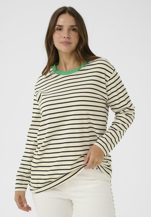 Kvinde iført en langærmet cremefarvet skjorte med sorte vandrette striber og en grøn krave, kombineret med hvide bukser mod en ensfarvet baggrund.