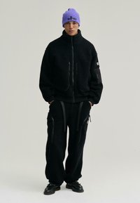 Czarna polarowa kurtka z zamkiem błyskawicznym i dwoma przednimi kieszeniami, w połączeniu z luźnymi czarnymi spodniami polarowymi z kieszeniami zapinanymi na zamek i czarnymi paskami. Fioletowa czapka.