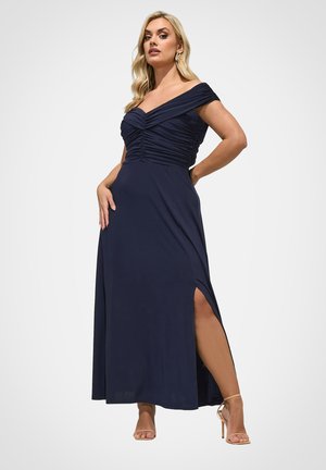 Kvinde poserer i en marineblå off-shoulder rynket kjole med højt slids, iført beige højhælede sandaler og gyldne øreringe i form af hoops.