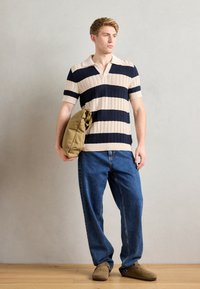 Tricou polo tricotat cu dungi orizontale bej și bleumarin, guler bej, asortat cu blugi largi albaștri și pantofi slip-on maro, ținând o geantă khaki.