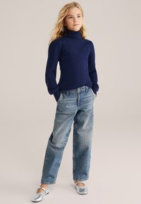 Sötétkék bordázott garbós pulóver hosszú ujjakkal, világoskék, laza fazonú farmernadrággal és ezüst lapos cipővel párosítva. Egyszínű háttér.