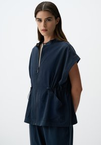 Gilet bleu marine en tissu léger, avec col montant, fermeture éclair, taille élastique et grandes poches avant.