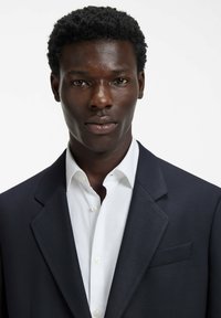 Granatowa, dopasowana marynarka o teksturowanej fakturze, noszona na białej koszuli zapinanej na guziki. Posiada klapy z wycięciem typu notch oraz jedną kieszeń na piersi.