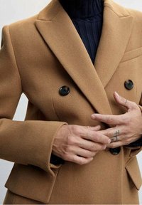 Cappotto cammello doppiopetto con bottoni neri e tessuto testurizzato, abbinato a un dolcevita blu navy a costine. Mani posizionate in vita.