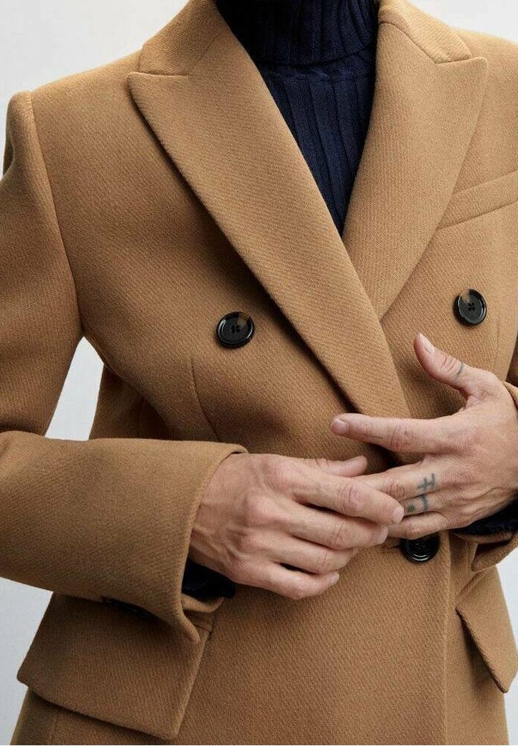 Cappotto cammello doppiopetto con bottoni neri e tessuto testurizzato, abbinato a un dolcevita blu navy a costine. Mani posizionate in vita.
