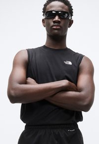 Μαύρο αμάνικο πουκάμισο με το λογότυπο "The North Face", φορεμένο με σκούρα γυαλιά ηλίου και μαύρες σορτς, με άνετη εφαρμογή και ομαλή υφή.