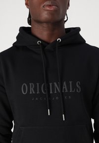 Sudadera negra con bolsillo frontal. Presenta un logo en relieve en gris oscuro, ojales metálicos en tono plateado y cordón. Tejido suave y forma clásica.