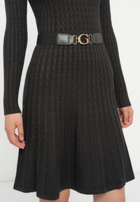 Abito lavorato a maglia nero con maniche lunghe e un corsetto aderente, che si allarga in una gonna. Accompagnato da una cintura nera con una fibbia dorata.