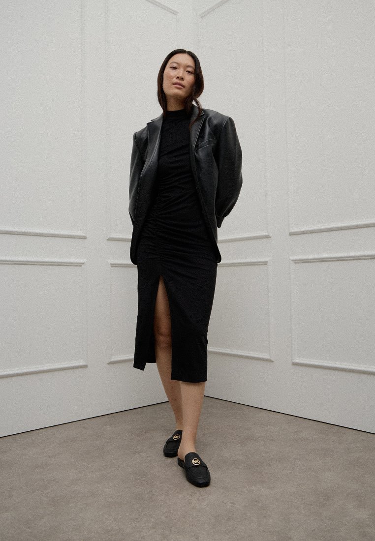 Zwarte ruche jurk met een split aan de zijkant, gecombineerd met een zwarte leren jas en loafers met gouden hardware accenten. Neutrale achtergrond.