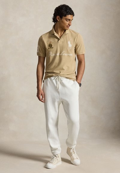 Άνδρας που φοράει μπεζ polo Ralph Lauren, λευκή φόρμα με κορδόνι και μπεζ αθλητικά παπούτσια, στέκεται με το ένα χέρι στην τσέπη.
