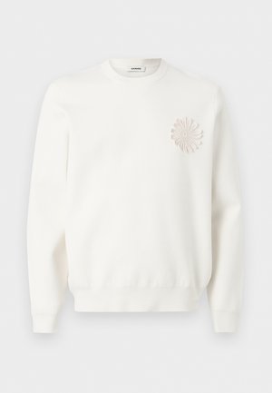 Jeune homme aux cheveux bouclés portant une tenue entièrement blanche : pull avec détail floral, pantalon large et chaussures beiges à enfiler, debout, main sur la hanche.