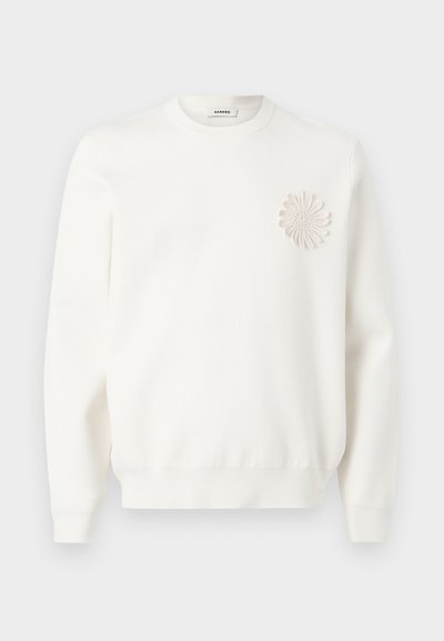 Λευκή μπλούζα με μακριά μανίκια, στρογγυλή λαιμόκοψη και υφασμάτινη κυκλική λουλουδένια εφαρμογή στο αριστερό στήθος.