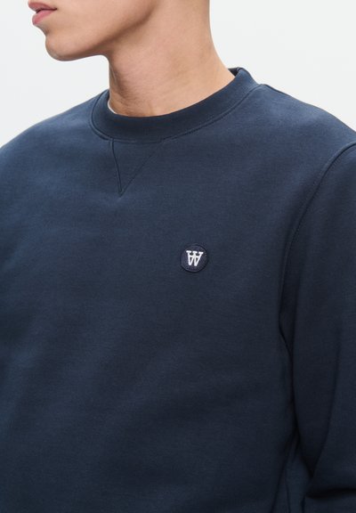 Sweat-shirt bleu marine à col rond, texture douce, et un petit logo circulaire sur la poitrine. Il présente une coupe classique et des poignets côtelés.