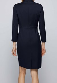 Námořnické modré šaty s dlouhými rukávy, strukturovaný design, zapínání na zip na zadní straně a délka do kolen. Mají přizpůsobený střih a jemné švy.