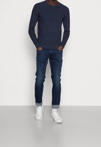 Granatowy sweter z dzianiny z okrągłym dekoltem, zestawiony z ciemnoniebieskimi dżinsami. Dopełnieniem stroju są białe trampki z okrągłym noskiem i podwiniętymi mankietami.